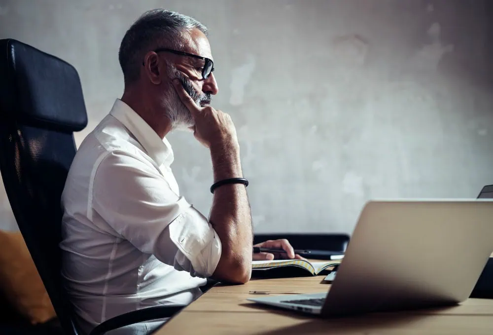 Adult bearded businessman wearing white shirt and working at the wood table in modern loft.Man making notes his notebook.Horizontal, blurred background