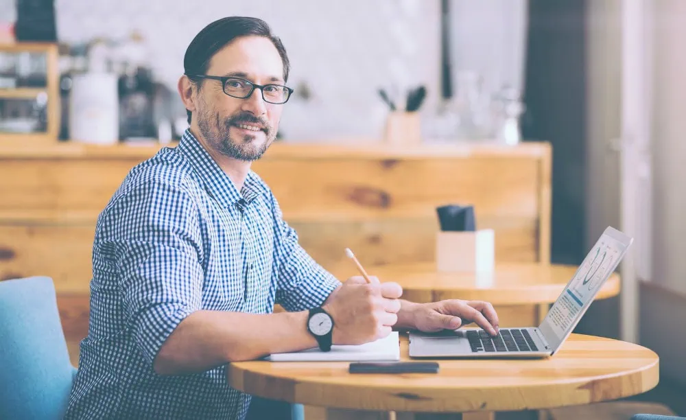 Business affairs. Pleasant smiling senior cafe owner managing his business and using laptop while sitting at the table